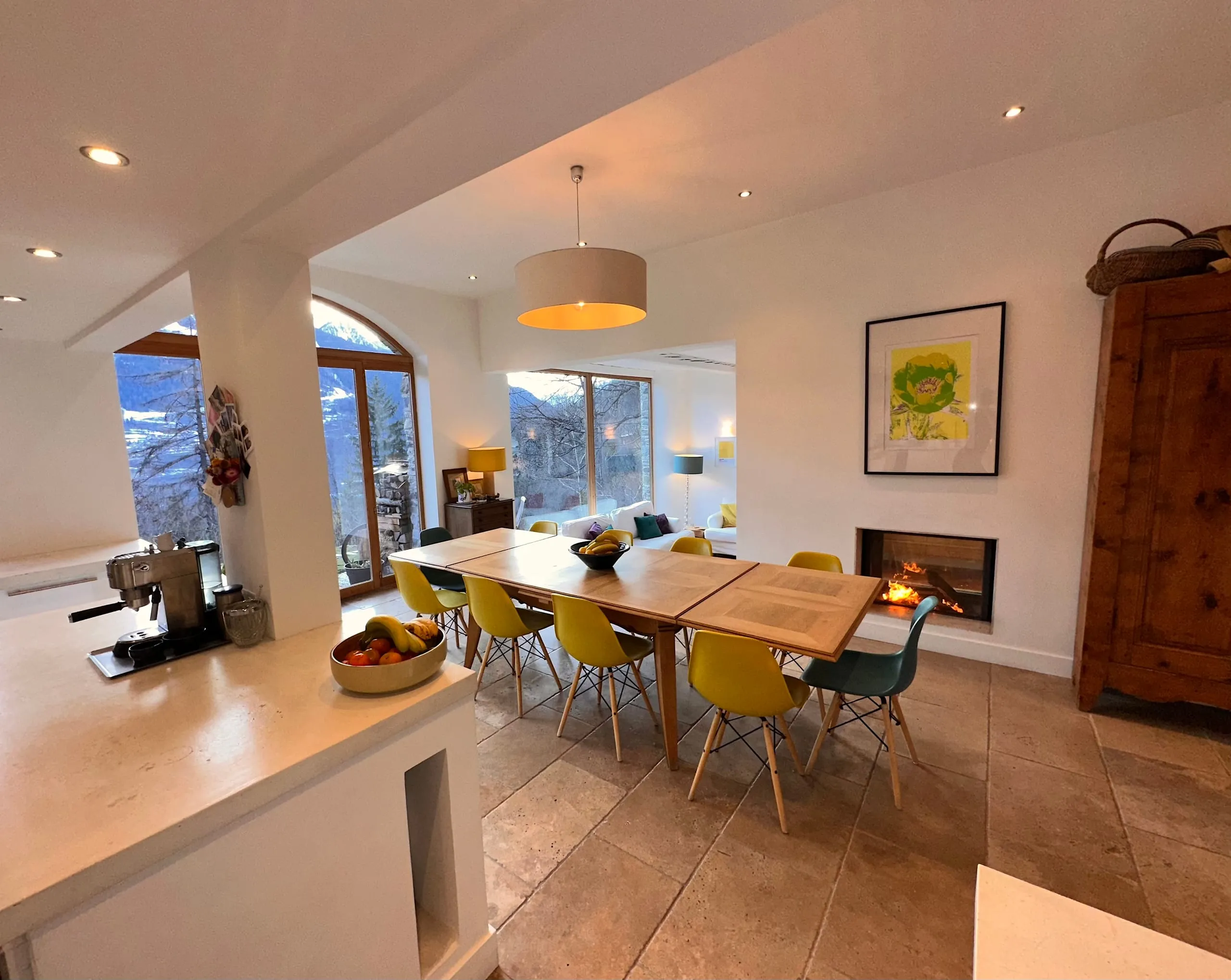 The dining area seen from the kitchen, the long table beside the fire, with the lounge and its windows beyond